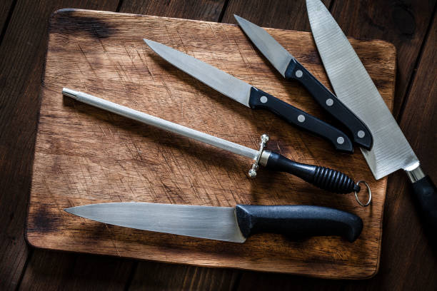 Sharpening steel and various kitchen knives on wooden cutting board Top view of a sharpening steel and various kitchen knives shot on wooden cutting board. Predominant color is brown. Low key DSRL studio photo taken with Canon EOS 5D Mk II and Canon EF 100mm f/2.8L Macro IS USM. knife sharpening stock pictures, royalty-free photos & images